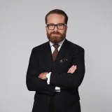 Maximilian Zirm mit dunkelblauem Anzug, brauner Krawatte und schwarzer Brille vor grauem Hintergrund.