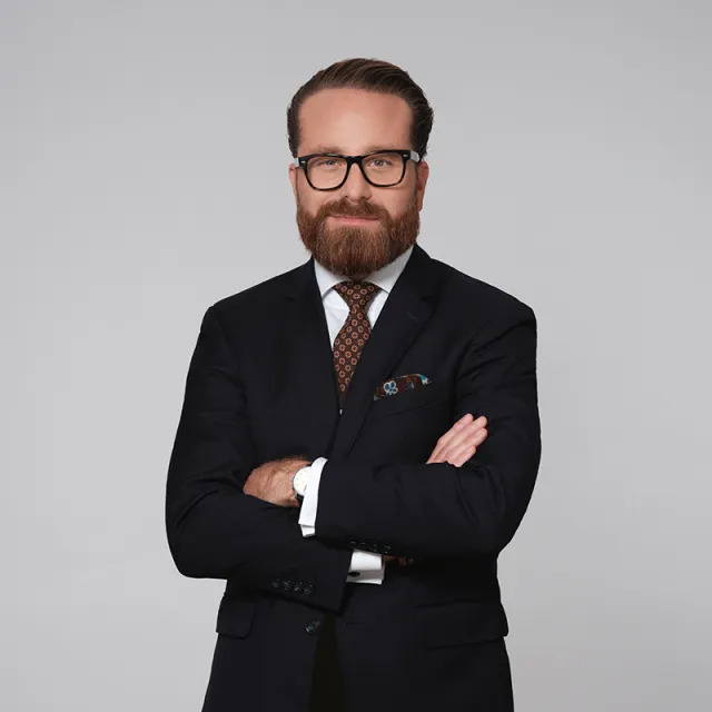 Maximilian Zirm mit dunkelblauem Anzug, brauner Krawatte und schwarzer Brille vor grauem Hintergrund.